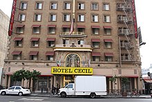 The lower five stories of a tan brick building in a city. Its lower windows have awnings; there is a fire escape on the right. At the street there is a large shelter over the entrance with «Hotel Cecil» on it in black type on a yellow background. A small truck and car, both white, are parked on the street in front.