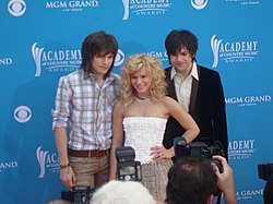 Two young men with a long dark hair and a young woman with long blonde hair posing for photographers in front of a blue backdrop