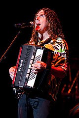 A photograph of «Weird Al» Yankovic, singing through a microphone and playing a harmonica.